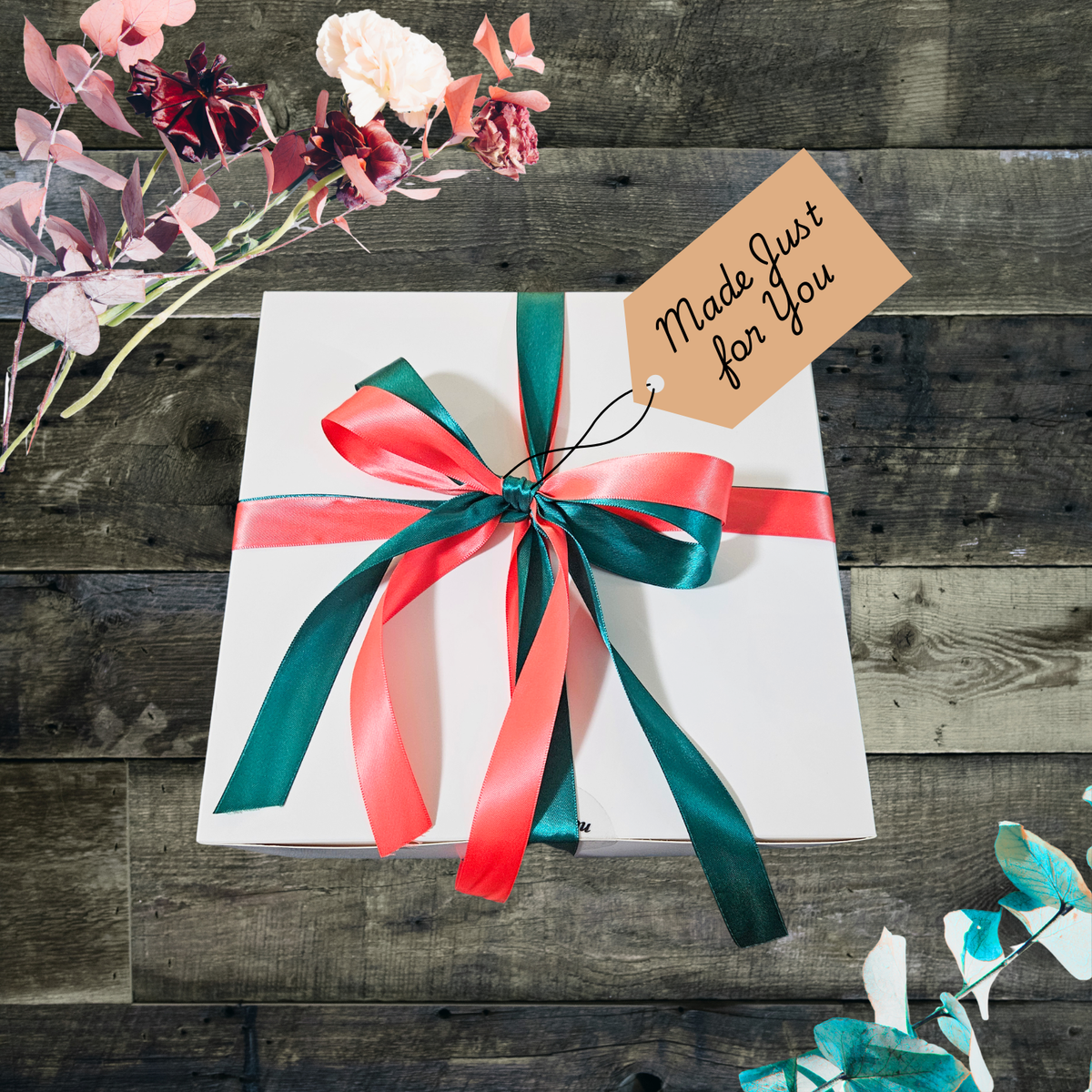 A white gift box tied with coral and dark teal ribbon, featuring a kraft paper tag reading 'Made Just for You,' set on a rustic wood surface with dried florals and eucalyptus.