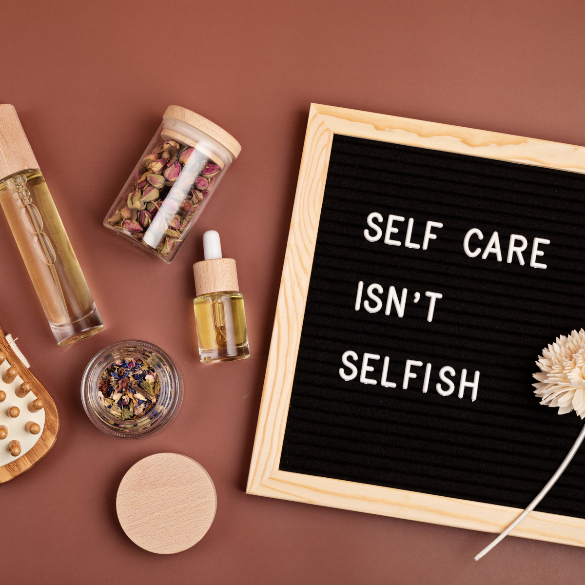 A flat‑lay arrangement of self‑care items surrounding a black felt letter board that reads “Self Care Isn’t Selfish,” including dried flowers in glass jars, a wooden massage brush, skincare bottles, and a beige flower on a warm brown background.