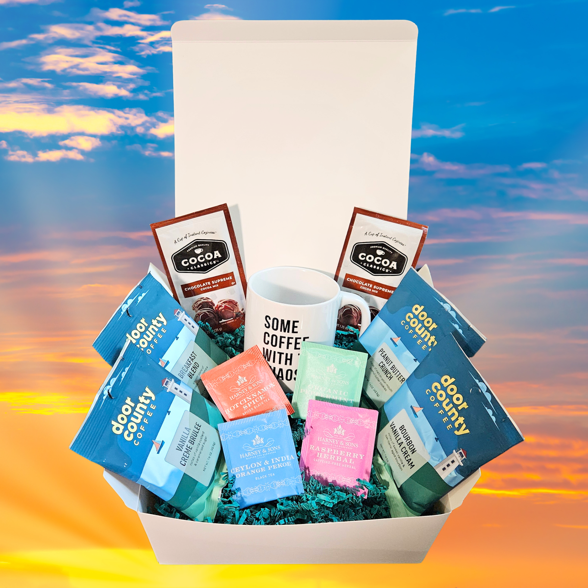 A curated warm beverage gift basket in a white box, featuring Door County flavored coffees, a white mug with black text, hot cocoa, and 4 different flavored tea bags, set against a sunset sky