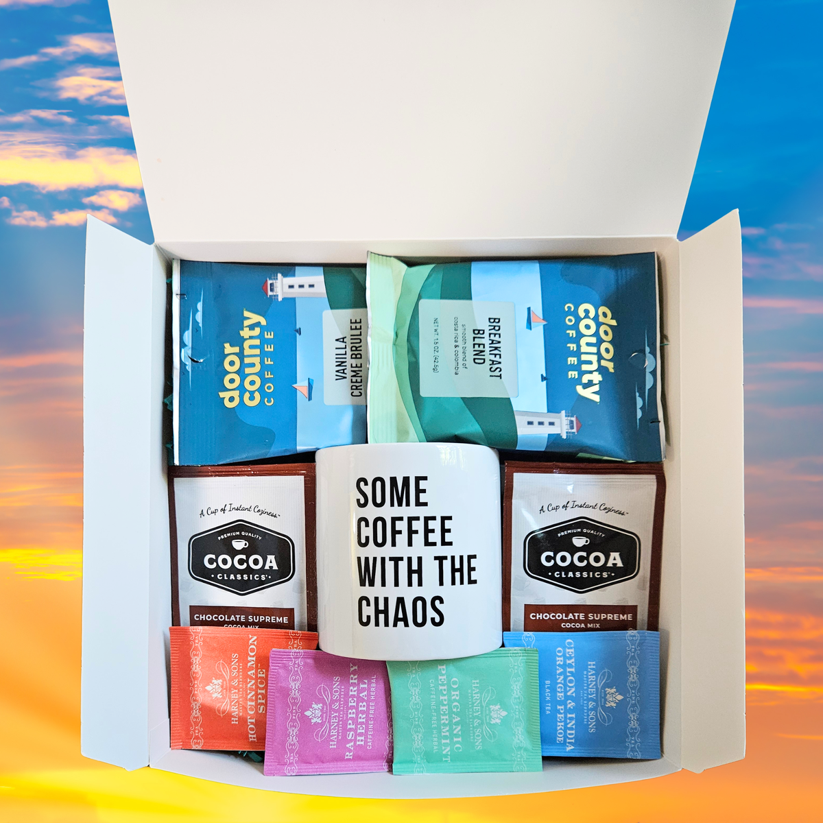 A curated warm beverage gift basket packed neatly in a white box, featuring Door County flavored coffees, a white mug with black text, hot cocoa, and 4 different flavored tea bags, set against a sunset sky