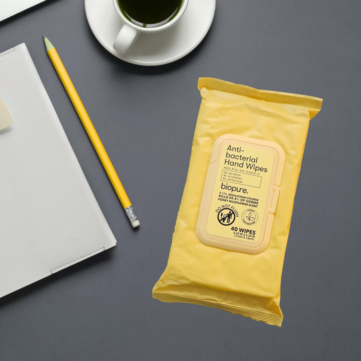 A yellow packet of Biopure Honey Wildflower scent Anti-Bacterial Hand Wipes is shown in a styled flat-lay setting on a dark gray surface, surrounded by a yellow pencil, a white notebook, and a cup of coffee.