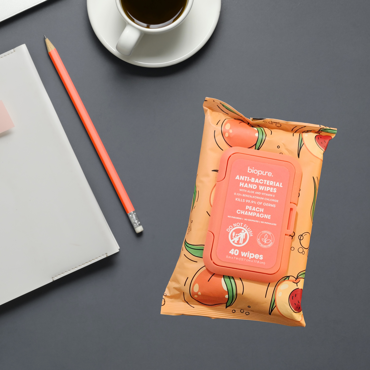A package of biopure. Anti-Bacterial Hand Wipes in Peach Champagne scent, styled on a dark gray desk next to a peach pencil, a white notebook, and a cup of coffee.