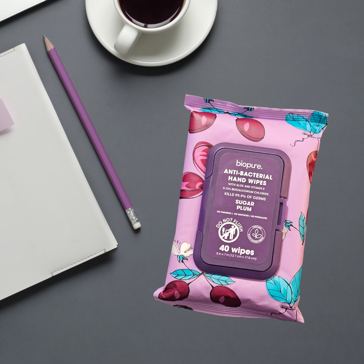 A package of biopure. Anti-Bacterial Hand Wipes in Sugar Plum scent, styled on a dark gray desk next to a purple pencil, a white notebook, and a cup of coffee.