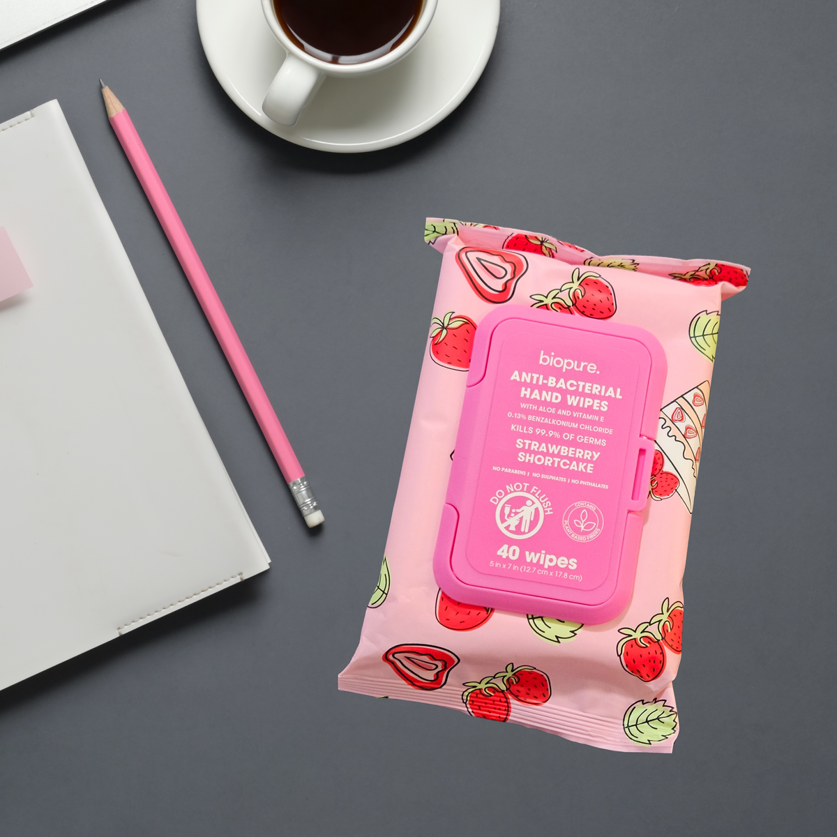 A package of biopure. Anti-Bacterial Hand Wipes in Strawberry Shortcake scent, styled on a dark gray desk next to a pink pencil, a white notebook, and a cup of coffee.