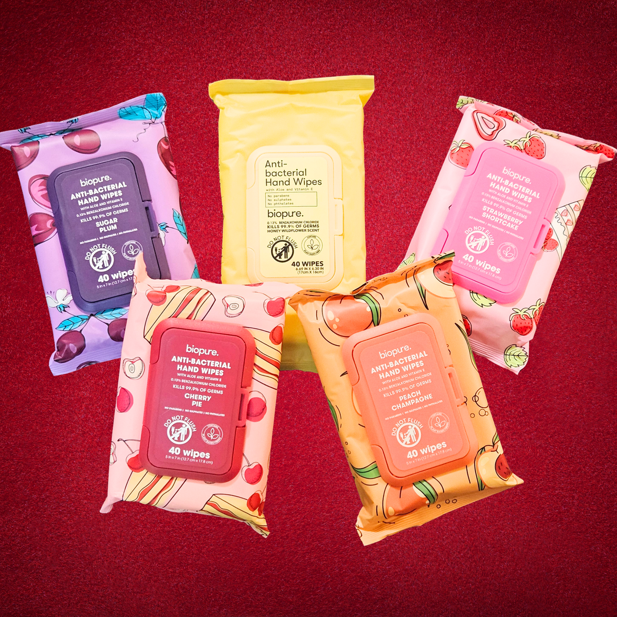 Five Biopure antibacterial hand wipes packages arranged on a dark red textured background. The packages feature different fruit and flavor designs in purple, yellow, pink, orange, and red, with the center yellow pack standing upright and the others fanned out around it.