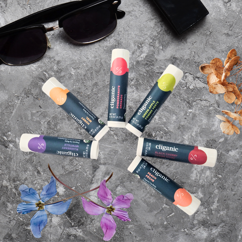 Six Cliganic organic lip balm tubes arranged in a circle on a textured gray surface, surrounded by sunglasses, a black wallet, dried leaves, and two pressed flowers for a styled, natural product display.