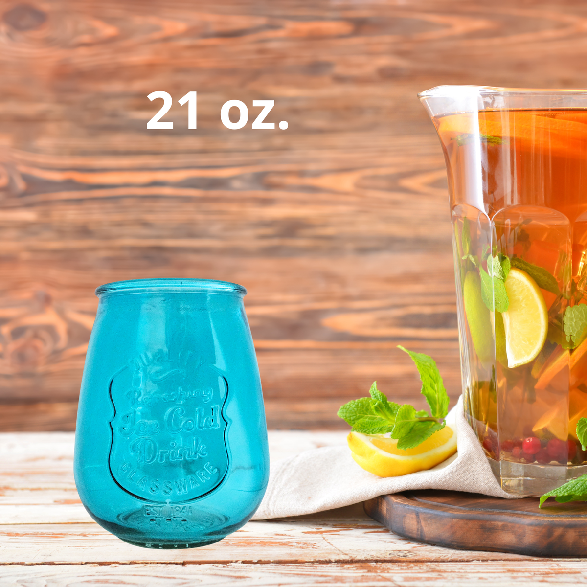 A 21 oz. teal blue glass drink tumbler shown next to a glass pitcher filled with iced tea, lemon slices, and mint, set on a rustic wooden table.