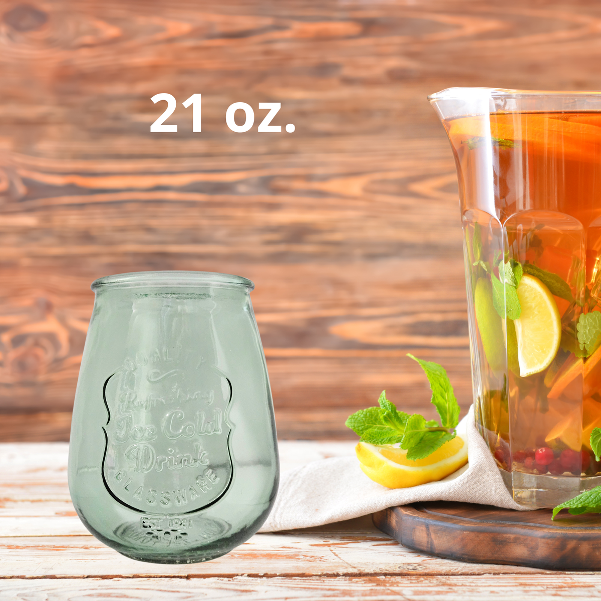 A 21 oz. green glass drink tumbler shown next to a glass pitcher filled with iced tea, lemon slices, and mint, set on a rustic wooden table.