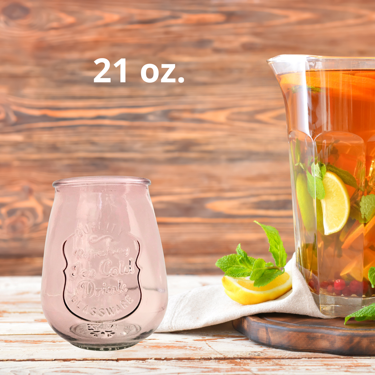 A 21 oz. pink glass drink tumbler shown next to a glass pitcher filled with iced tea, lemon slices, and mint, set on a rustic wooden table.