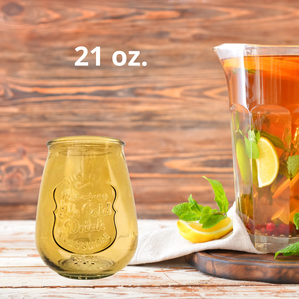 A 21 oz. yellow glass drink tumbler shown next to a glass pitcher filled with iced tea, lemon slices, and mint, set on a rustic wooden table.