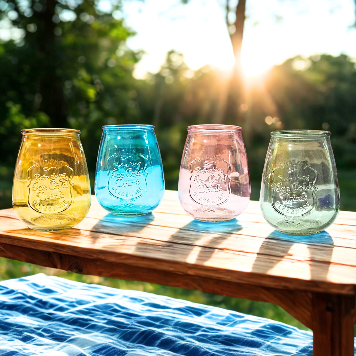 Four embossed glass mason-style cups in yellow, blue, pink, and green, lined up on a wooden table outdoors with a blurred forest background and bright sunlight.