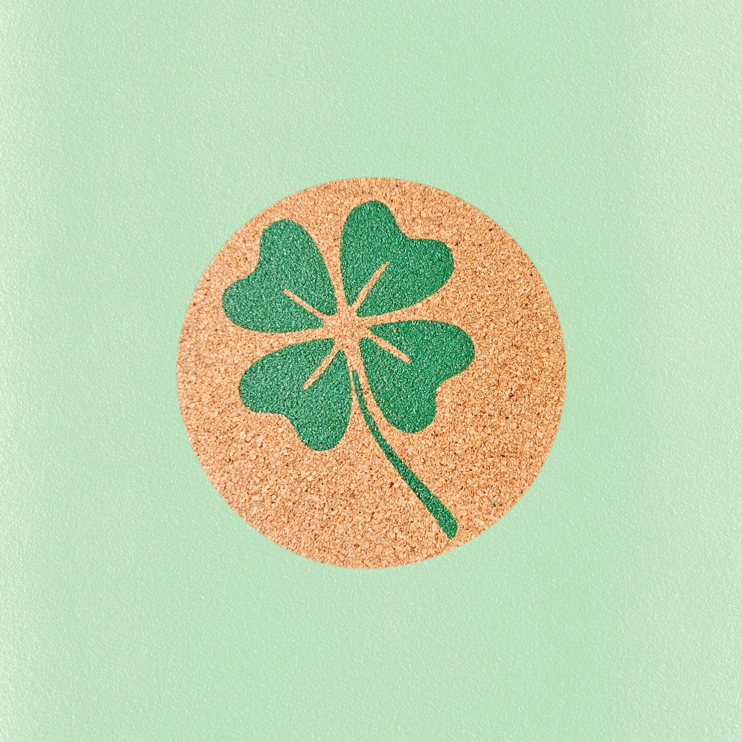 A round cork coaster featuring a green four-leaf clover design, centered on a solid mint green background.