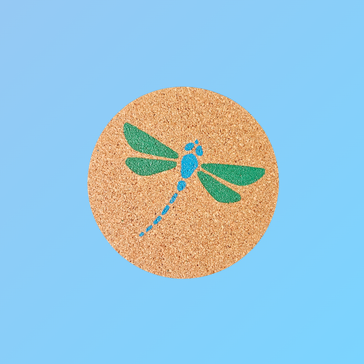 A round cork coaster featuring a green and blue dragonfly design, centered on a solid light blue background.