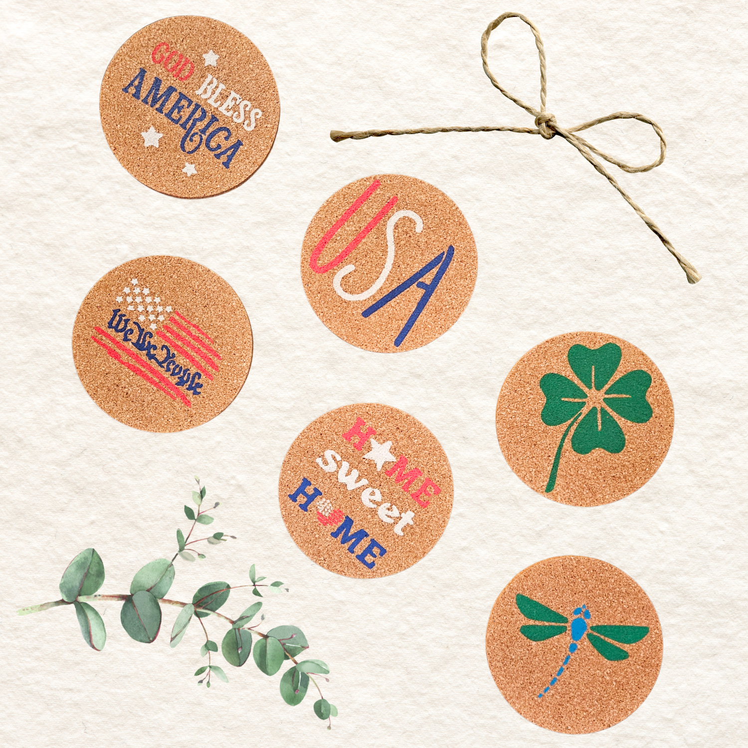 Six round cork coasters with patriotic and American-themed designs, including 'God Bless America,' 'USA,' 'I Love This Country,' 'It's So Sweet Home,' a green four-leaf clover, and a blue & green dragonfly. The coasters are arranged on a textured white background with decorative eucalyptus stems and a piece of twine.