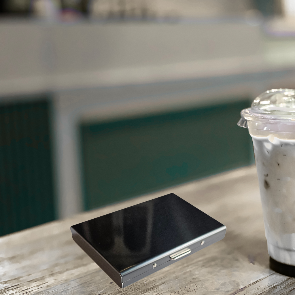 Closed black stainless steel RFID credit card holder on a wooden counter next to an iced coffee with blurred background.