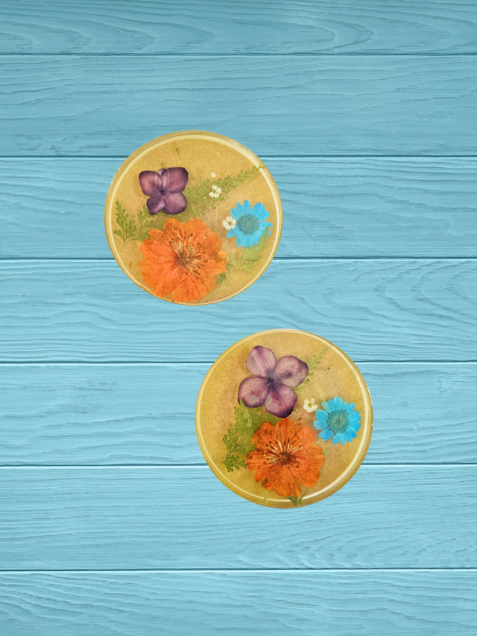 Two resin coasters with real pressed flowers in orange, blue, purple, and white on a light blue background.