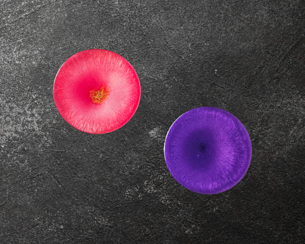 Two handmade resin coasters in pink and purple with radiant crystal‑burst patterns on a dark background.