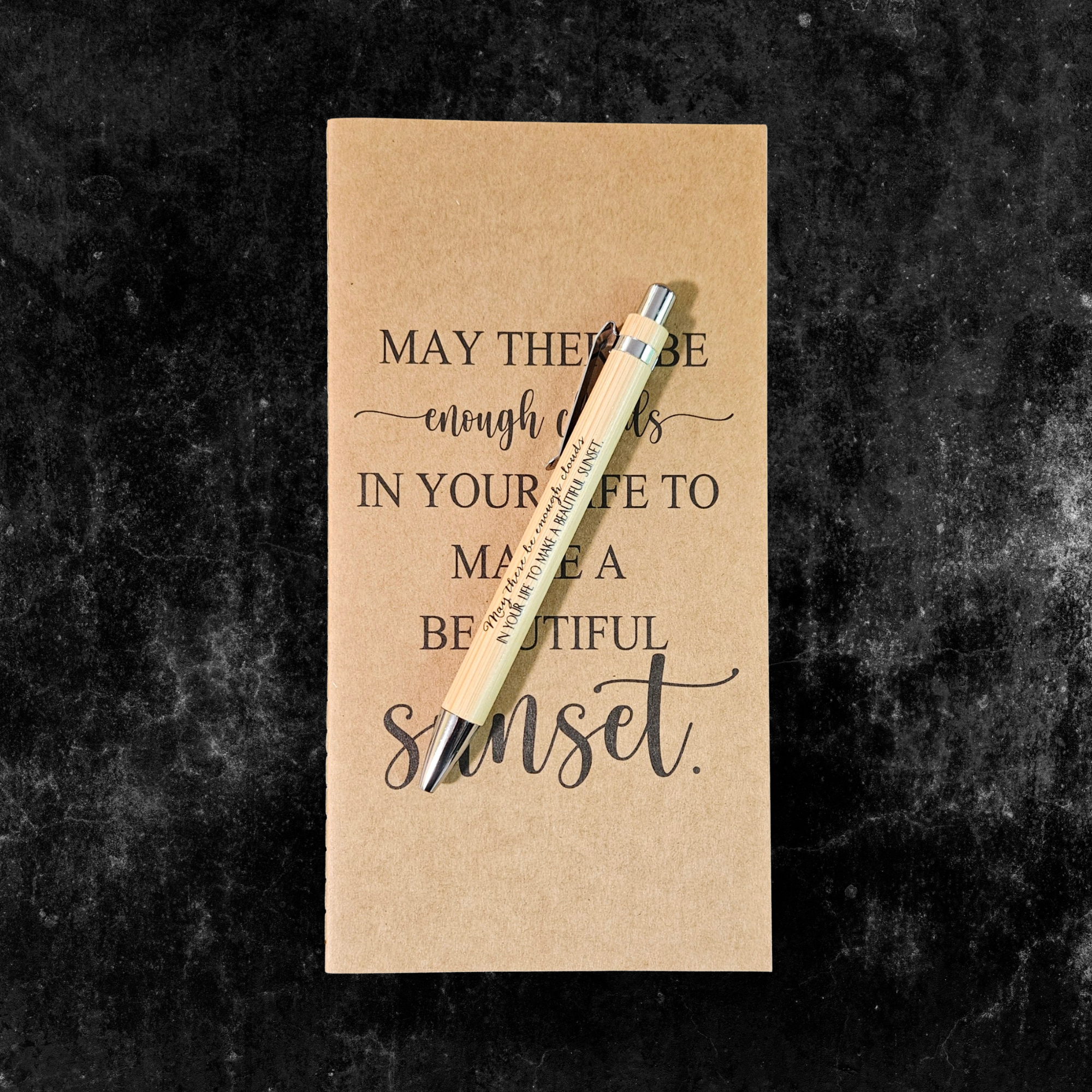 Kraft planner with motivational quote, May there be enough clouds in your life to make a beautiful sunset, and pen on a dark textured background
