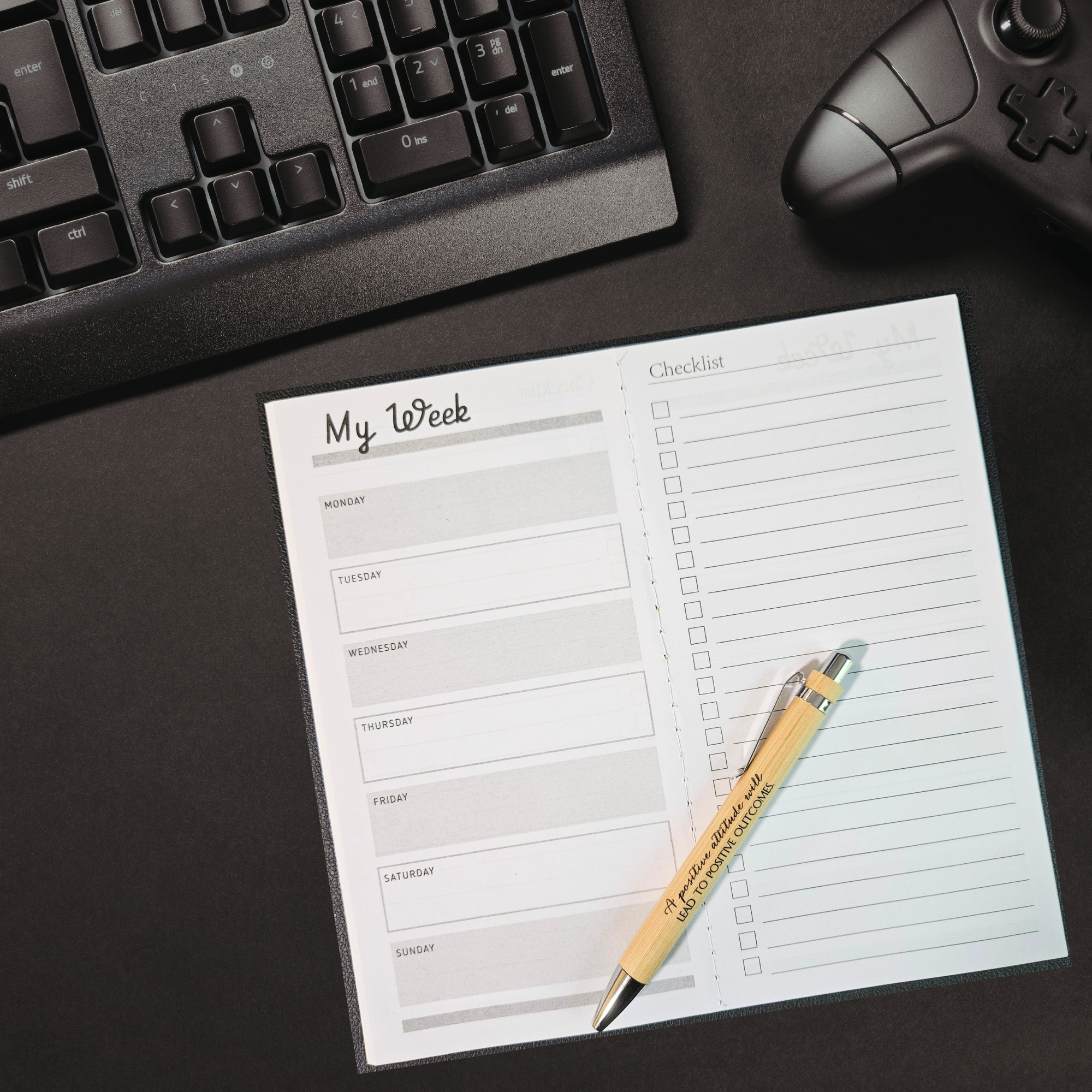Weekly planner opened on a desk with a bamboo pen, next to a computer keyboard & gaming controller.